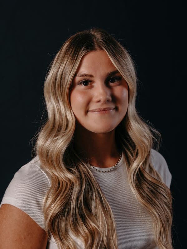 A woman with long, wavy blonde hair smiles softly against a dark background, wearing a white top and a beaded necklace.