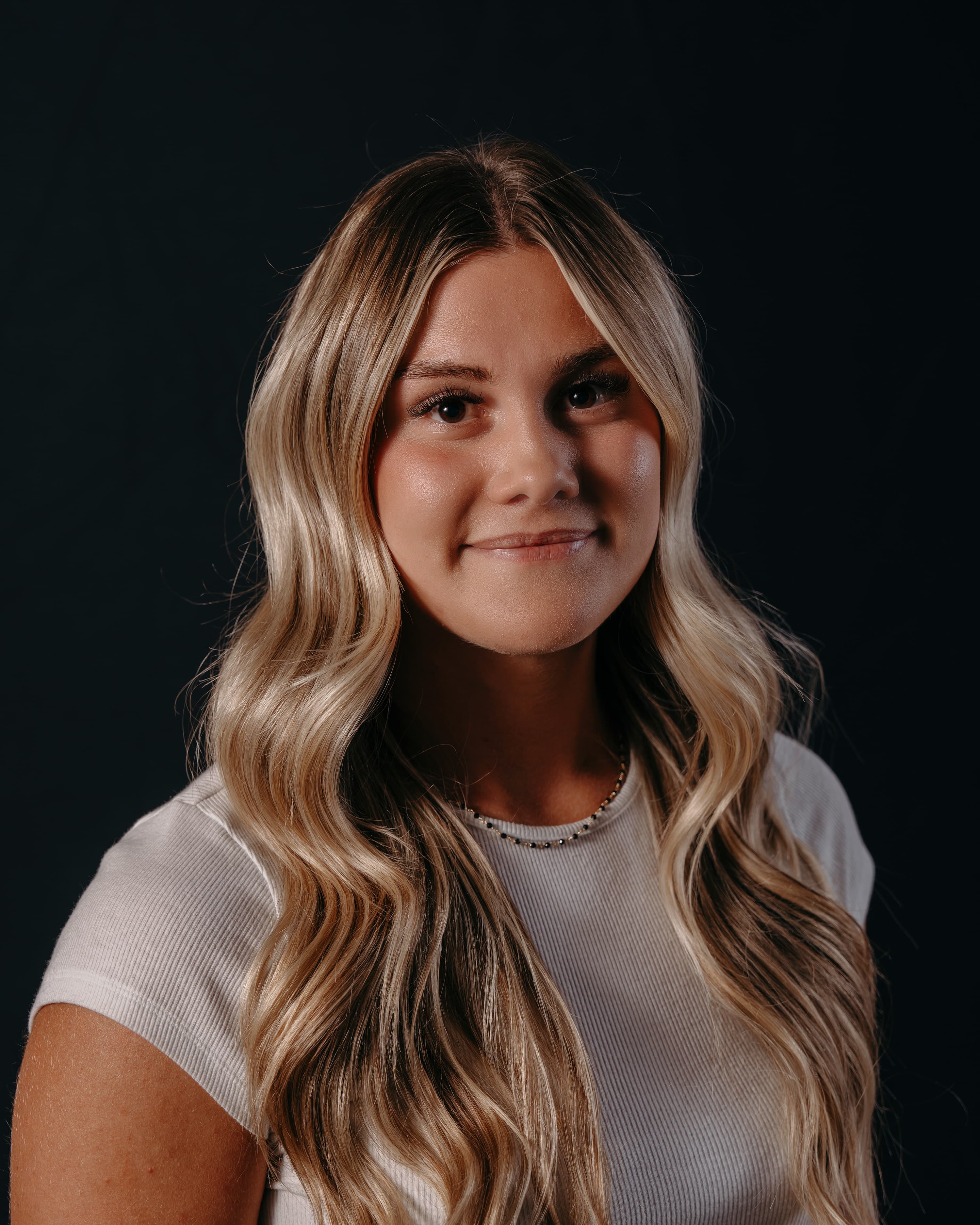 A woman with long, wavy blonde hair smiles softly against a dark background, wearing a white top and a beaded necklace.